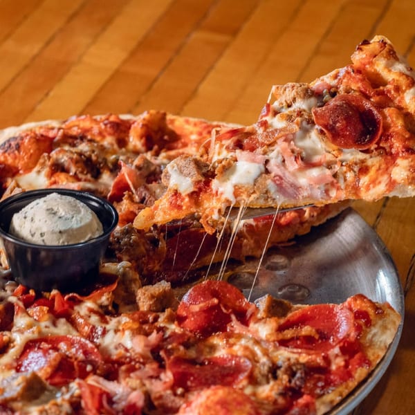 A slice of meat lover's pizza being lifted, with cheese stretching, next to a small cup of dip.