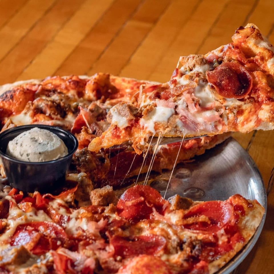 A slice of meat lover's pizza being lifted, with cheese stretching, next to a small cup of dip.