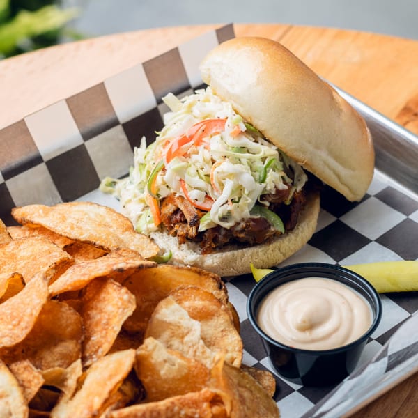 Pulled pork sandwich with coleslaw, a side of potato chips, and dipping sauce on a checkered tray.